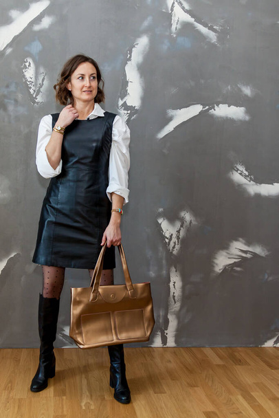 Fashionable and stylish girl posing in a black leather dress and a white shirt against a gray wall - Fotografie, Obrázek