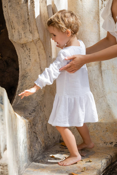 Side view of woman touching toddler daughter in summer dress on stone bench in Valencia - 写真・画像