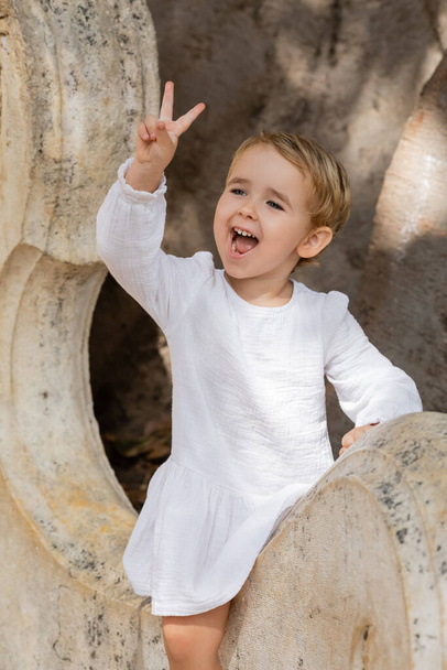 Excited toddler girl in dress showing peace sign on stone bench in Valencia  - Photo, Image
