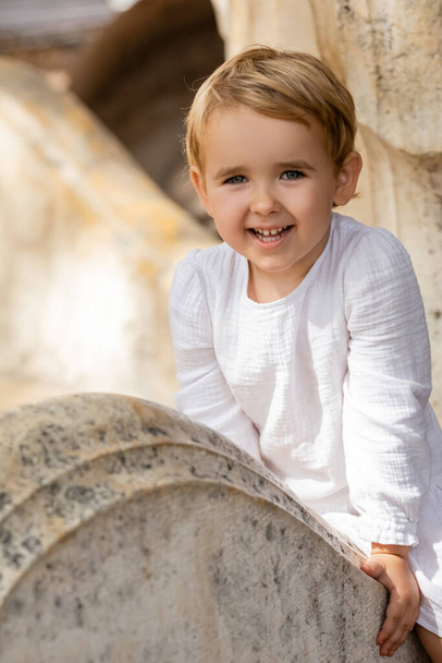 Mosolygó kislány fehér ruhában ül kő padon a parkban  - Fotó, kép