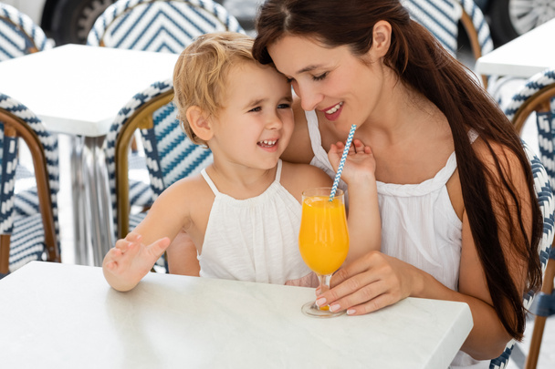 Женщина обнимает маленькую дочь возле апельсинового сока на столе в кафе на открытом воздухе в Валенсии  - Фото, изображение