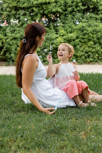Cheerful baby girl holding soap bubbles near mom on glass in park  - Photo, image