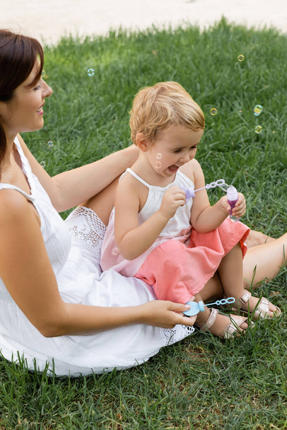 Happy mother looking at baby daughter playing with soap bubbles on lawn  - Foto, Imagen