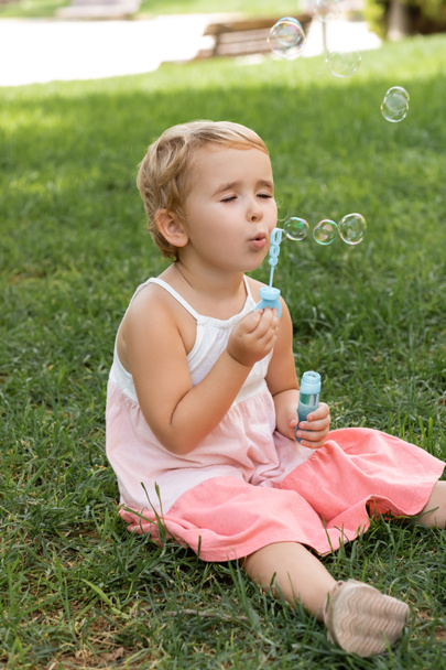 Portrait of toddler kid in dress blowing soap bubbles on lawn  - Фото, изображение