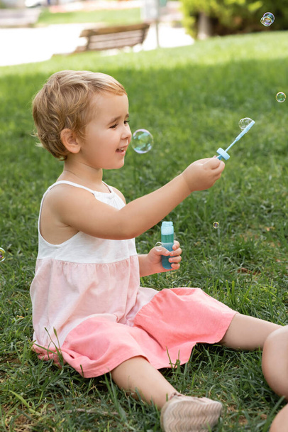 Cheerful child in summer dress playing with soap bubbles in park  - Photo, Image