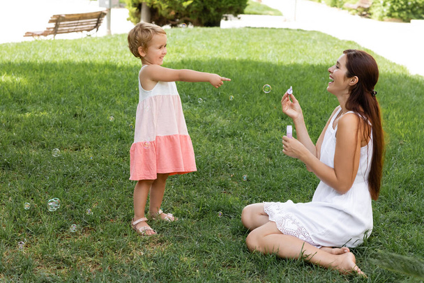 Улыбающаяся девочка указывает на маму с мыльными пузырями в парке  - Фото, изображение