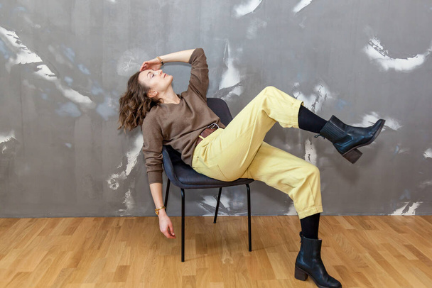 Fashionable and stylish girl posing in yellow trousers and a brown sweater against a gray wall - Foto, afbeelding