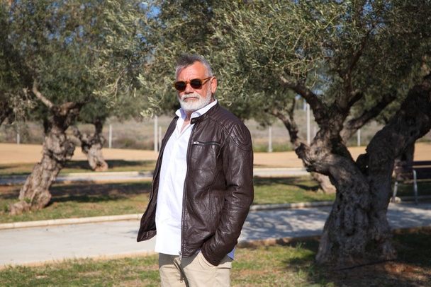 handsome mature man with beard and grey hair is walking in the park. The man is dressed in beige trousers, white shirt and leather jacket. Old age concept - 写真・画像