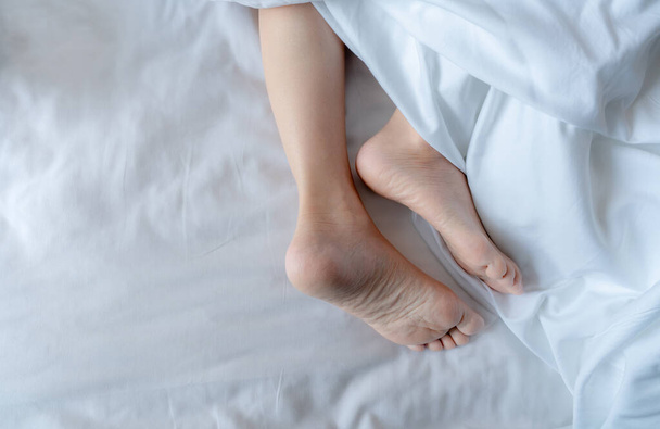 Woman barefoot on bed under white linen blanket in hotel or home bedroom. Healthy sleep and relaxation concept. Lazy Sunday morning. Bare feet of woman chilling sleep on white comfort bed and duvet. - Foto, afbeelding