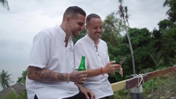 Happy laughing male gay couple having fun drinking beer and watching funny videos on phone at tropical island during vacation - 4K Horizontal video - Video, Çekim