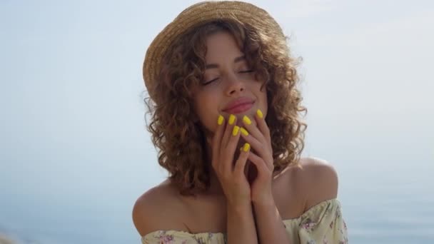 Seductive woman closed eyes enjoy sunlight on beautiful seacoast close up. Portrait of attractive curly girl wearing straw hat gentle touching face. Relaxed young lady posing on sunny summer beach. - Video