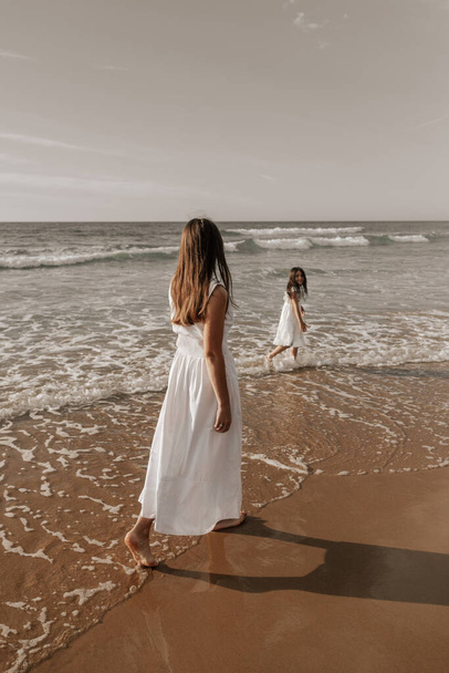 Barefoot mom in white dress with long hair walking after daughter while spending time on sandy beach near waving sea - Foto, Bild