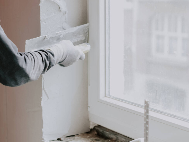 The hand of a guy in a gray T-shirt with sleeves applies fresh putty with a small spatula to the window opening, side view close-up. The concept of repairing apartments and houses, plastering walls, plastering services, at home. - Fotoğraf, Görsel