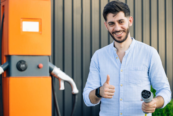 Male holds a charging cable in his hands. Caucasian bearded hipster man standing near electric charging station looking at camera and showing thumb up. Eco-friendly modern green transport. - 写真・画像