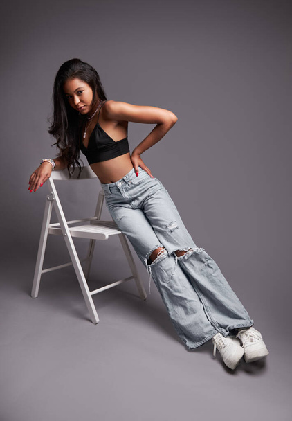 Full body trendy young Latin American woman in jeans and bra with hand on waist doing side plank, on chair and looking at camera against gray background - Foto, immagini