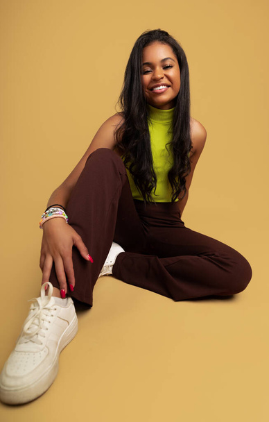 Full body of confident young Hispanic female teenager with long dark hair in trendy clothes, and sneakers sitting on floor and looking at camera against beige background - Zdjęcie, obraz