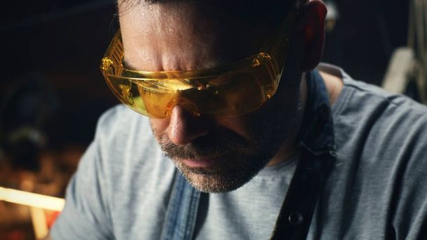 En el garaje, un hombre en ropa de trabajo y gafas está ocupado procesando metal para una parrilla de barbacoa por sí mismo para pequeñas empresas - Foto, imagen