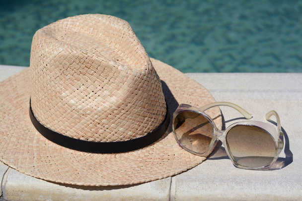 Stylish hat and sunglasses near outdoor swimming pool on sunny day, closeup. Beach accessories - 写真・画像