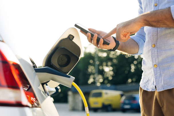 Male attaching in power cord to electric car using app on smartphone. Man plugging electric car from charging station. Businessman charging electric car on charging station at sunset - Foto, Imagen