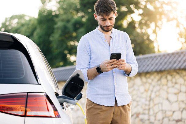 Caucasian bearded man using smart phone and waiting power supply connect to electric vehicles for charging the battery in car. Plug charging an Electric car from charging station. - Photo, Image