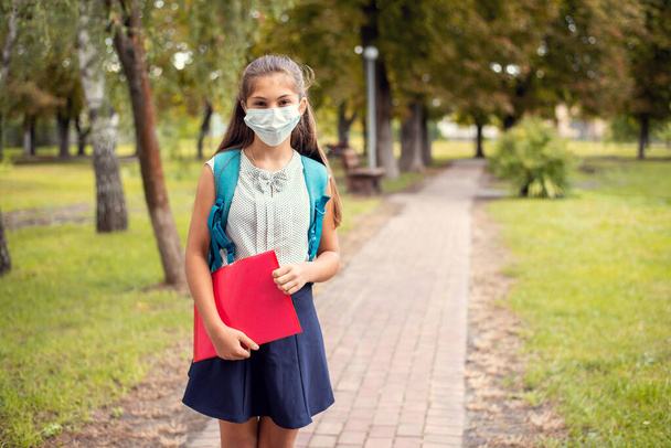 Школьница в защитной медицинской маске, носит рюкзак и держит красную папку с материалами, идущими в школу через парк - Фото, изображение