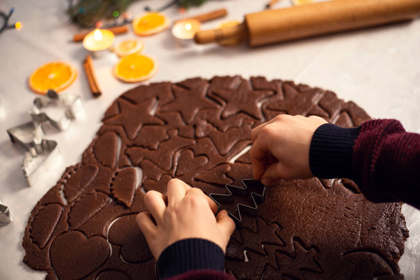 Eine Ruhe aus braunem Kakaoteig auf dem Tisch mit weihnachtlichem Dekor und Backzubehör. Die Hände eines Jungen, der Kekse in verschiedenen Formen herstellt. - Foto, Bild