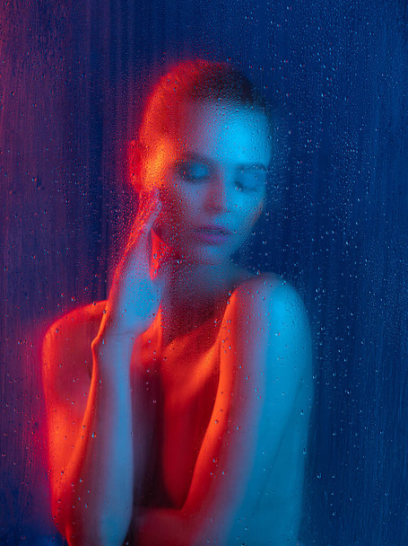 Portrait de jeune femme tendre avec un maquillage lumineux posant isolé sur fond bleu dans la lumière néon. Effet verre fantaisie. Concept de beauté, mode, style, cosmétologie, cosmétique. Espace de copie pour la publicité - Photo, image