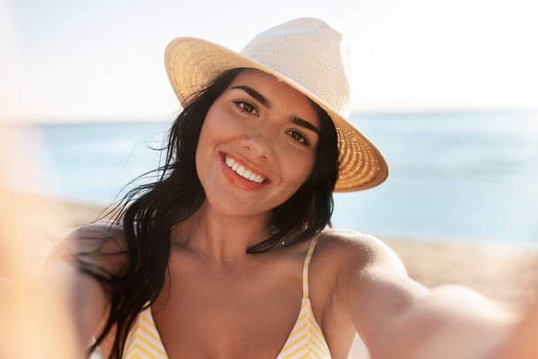 people, summer and swimwear concept - happy smiling young woman in bikini swimsuit and straw hat taking selfie on beach - Zdjęcie, obraz