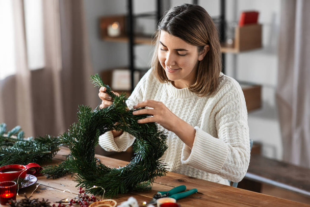 winter holidays, diy and hobby concept - happy smiling woman with fir branches making christmas wreath at home - Foto, afbeelding