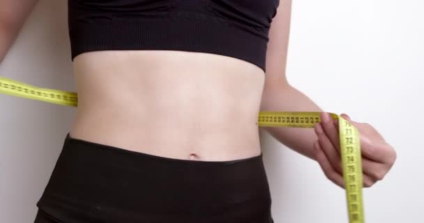 A young woman measures her waist with a tape while looking in the mirror. Beautiful female body. Healthy food diet and care concept. - Metraje, vídeo