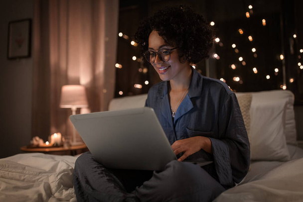 Technologie, Schlaf- und Ruhekonzept - glücklich lächelnde Frau im Schlafanzug mit Laptop, der nachts im Bett arbeitet - Foto, Bild