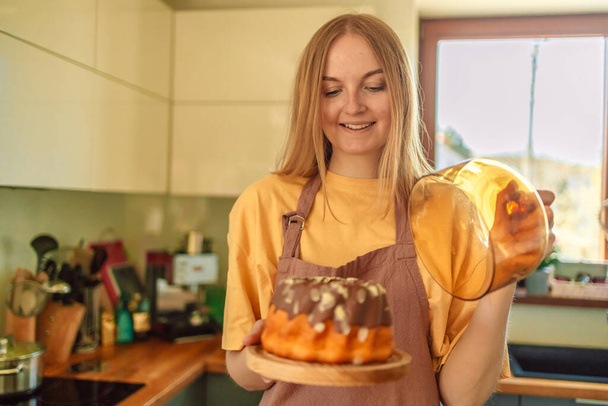 Uśmiechnięta młoda blondynka kucharz kucharz w fartuchu stoi w kuchni, pokazując smaczne ciasto.  - Zdjęcie, obraz