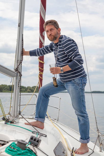 Businessman with a beard in a striped sweater stands on the nose of a white yacht and drinks whiskey from a glas - 写真・画像