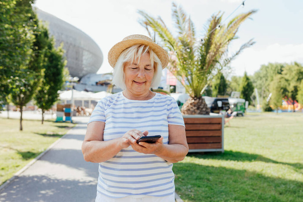 Mature woman in casual summer clothes with straw hat browsing phone while wandering streets of city. Caucasian blogger holding cellphone pondering thinking on idea for social publication outdoors - Valokuva, kuva