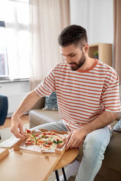 food delivery, consumption and people concept - happy man opening box and eating takeaway pizza at home - Foto, afbeelding