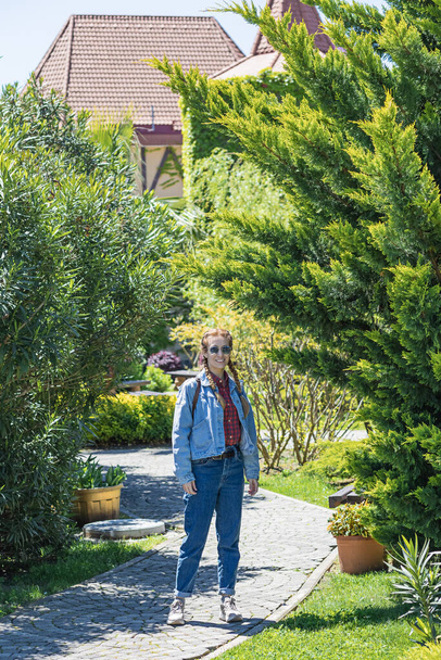 Eine lächelnde Frau im Jeans-Outfit steht an einem sonnigen Frühlingstag auf einem gepflasterten Weg zwischen Parkbäumen.. - Foto, Bild