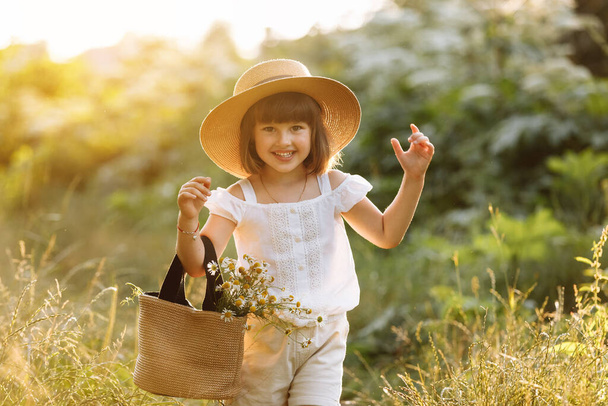Portrait of smiling little girl walks in the rays of a sunset in a flowering meadow, enjoying the summer, warmth, flowers, freedom. Child with straw hat and bag is having fun outdoors. - Foto, Bild