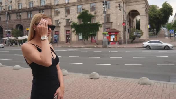 A young blonde woman in a black dress walks near the road in the capital. Portrait of a blonde woman in sunglasses. Woman walking in the city - Materiał filmowy, wideo