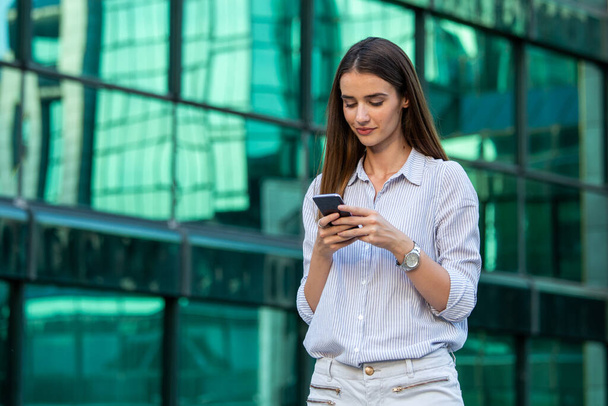 Geschäftsfrau im Gespräch auf dem Handy auf der Straße mit Bürogebäuden im Hintergrund. Junge Frau mit Smartphone steht vor verschwommenem Gebäude. - Foto, Bild