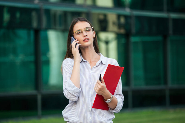 Businesswoman in formal wear having trouble with contracts talking on the phone and holding clipboard. Businesswoman angry abount about paperwork failure at workplace, executives having conflict - 写真・画像