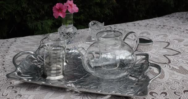 Erwachsene weibliche Hände entfernen den Deckel und das Ei einer gläsernen Teekanne, die auf einem Metalltablett steht, zwei Tassen, einen Tisch mit einer Vase mit rosa Blüten und eine dünne braune gestrickte Tischdecke. Gartentee-Zeitkonzept - Filmmaterial, Video