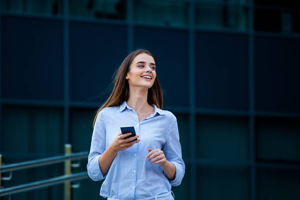 Ευτυχισμένο χαμογελαστή γυναίκα επιχείρηση μιλά σε κινητό smartphone στο δρόμο με κτίρια γραφείων στο παρασκήνιο. - Φωτογραφία, εικόνα