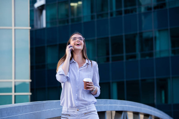 Gelukkig lachende zakenvrouw praten op mobiele smartphone en koffie drinken uit wegwerp papier Cup in de straat met kantoorgebouwen op de achtergrond. - Foto, afbeelding