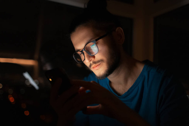 Night portrait of serious man looking in smartphone on background of window. - Foto, imagen