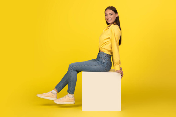 Vidám Y-generációs nő mosolyog a kamerába ül a kockán pózol sárga háttér felett. A Boldog Nő Stúdiójában. Szépség és stílus. Teljes hosszúságú lövés - Fotó, kép