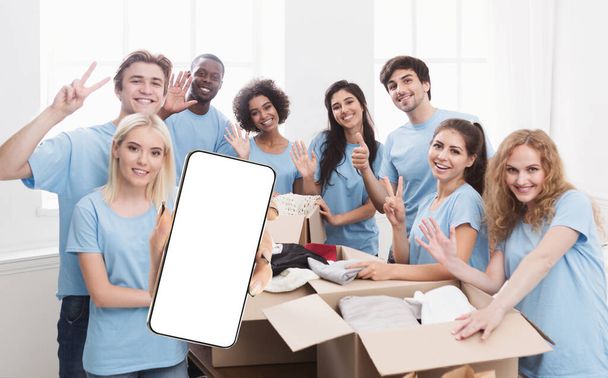 Volunteering App. Multiethnic Group Of Volunteers Showing Big Blank Smartphone While Sorting Clothes, Happy Young People Showing Mobile Phone With White Screen, Recommending Application, Mockup - Photo, image