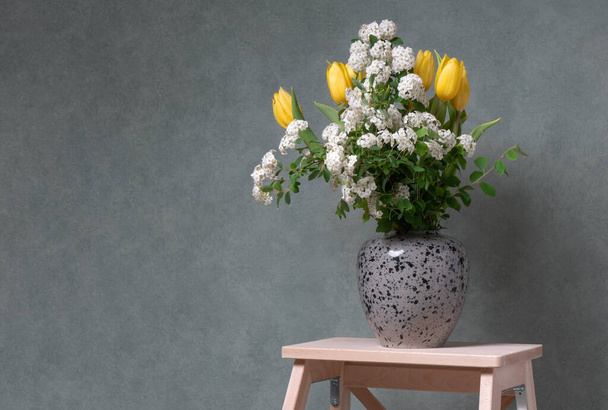 Spring bouquet of flowers in a gray vase on a gray background on a wooden staircase. Yellow tulips and white flowers. Fresh and colorful flowers. Vksna aroma. Joyful mood in the house. Comfort and tranquility. - Photo, Image
