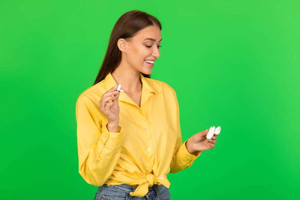 Happy Woman Holding Earbuds Listening To Music Standing Over Green Background. Studio Shot Of Cheerful Lady Posing With Earphones. Musical Application Concept - 写真・画像