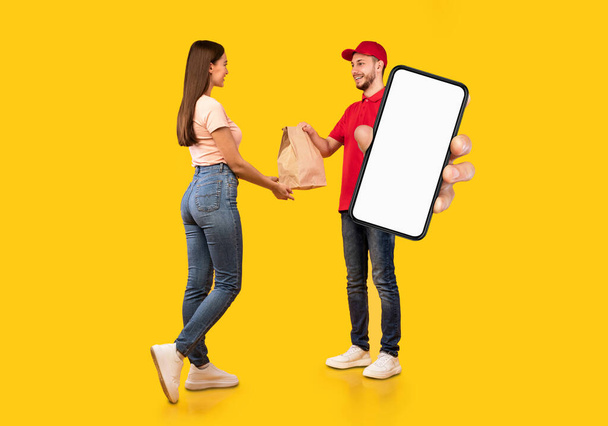 Delivery App. Male Courier Holding Big Blank Smartphone And Giving Package To Female Customer, Recommending Application Or Website, Standing Together Over Yellow Background In Studio, Mockup, Collage - Valokuva, kuva