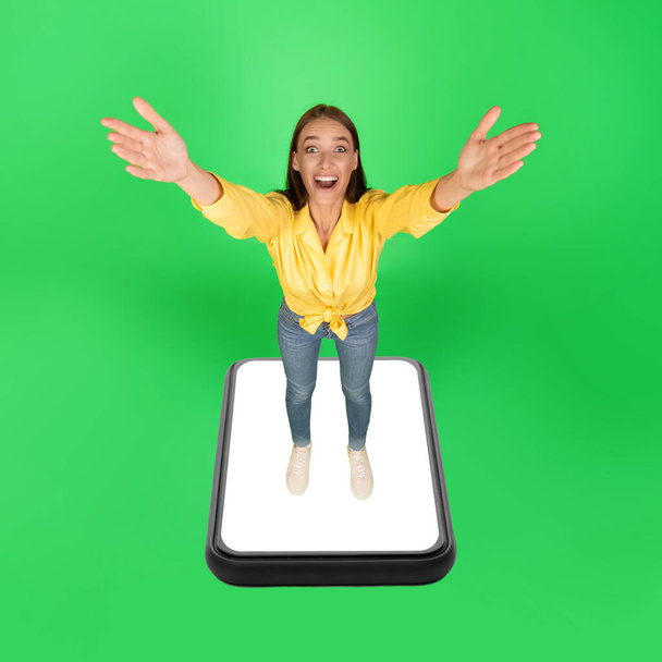 Aufgeregte Frau, die auf riesigem Smartphone-Bildschirm steht und die Arme zur Kamera ausstreckt, posiert vor grünem Hintergrund im Studio. Große mobile Anwendung. Collage, quadratische Aufnahme - Foto, Bild